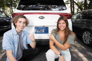 two students kneel at the trunk of a car with the Argos license plate